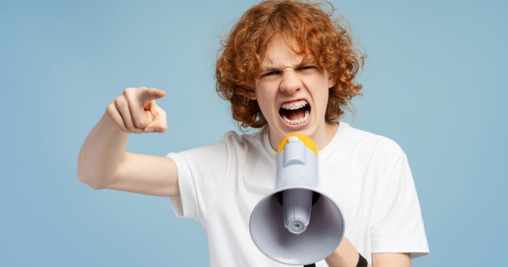 Nervous red haired curly teenager with braces wearing white t shirt holding loudspeaker looking at camera isolated on blue background. Concept of announcement, communication