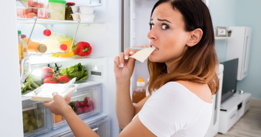 Young Woman Eating Slice Of Cheese In Front Of Open Refrigerator In Kitchen
