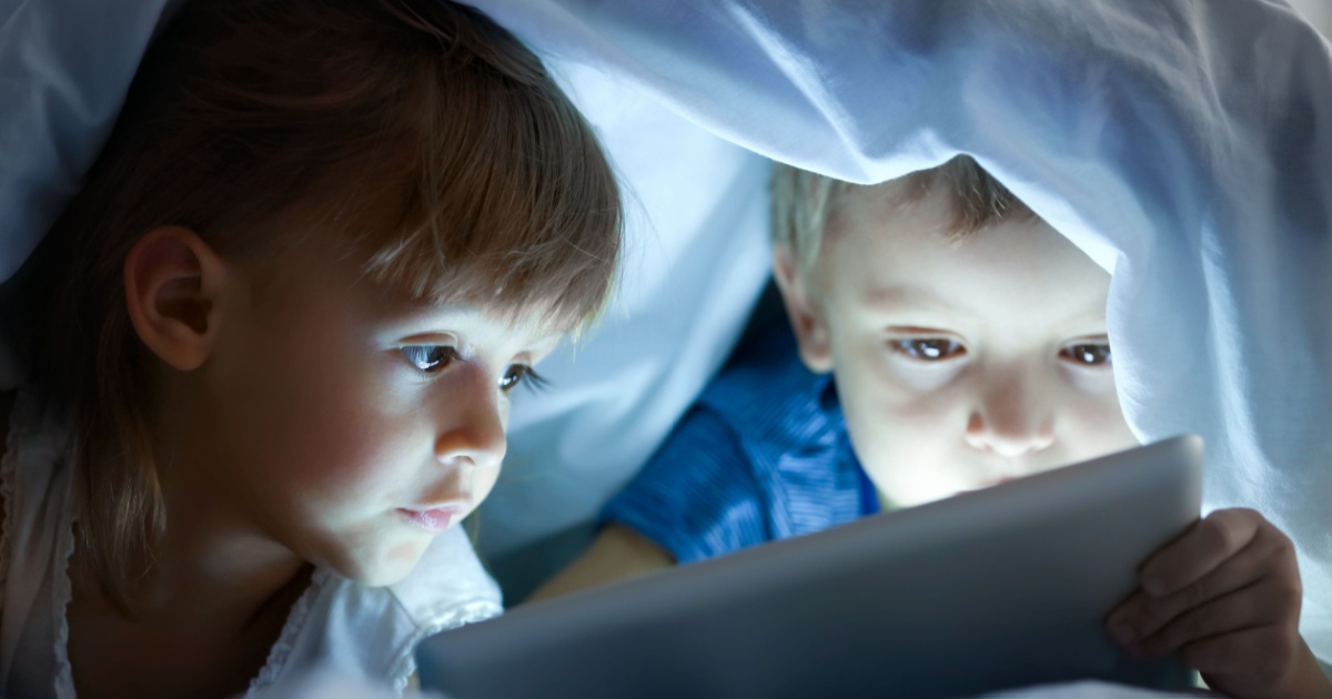 Cute little children lying under blanket with tablet computer