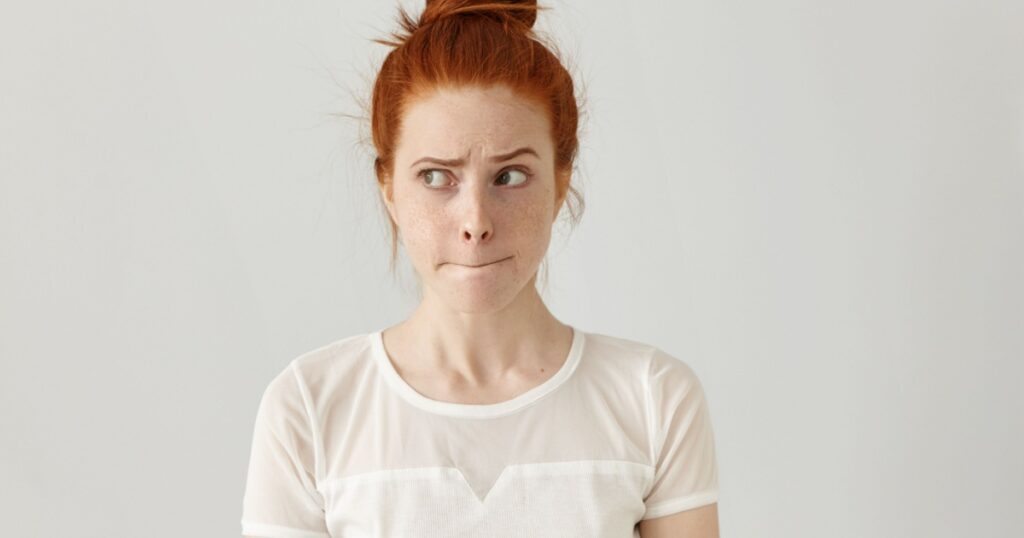 Indoor shot of cute redhead girl looking away, having doubtful and indecisive face expression, pursuing her lips as if forbidden to say anything. Confused young female posing isolated at white wall