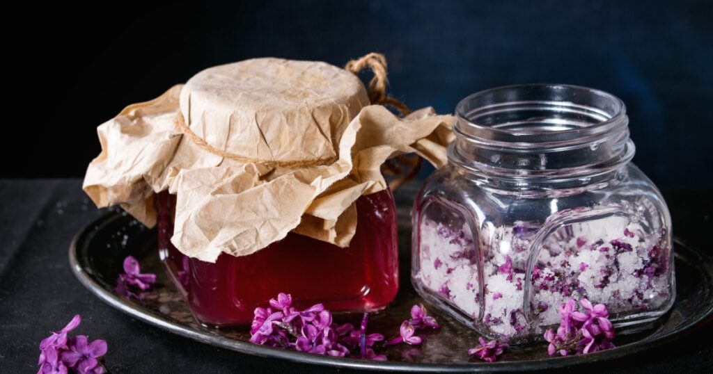 Glass jar of homemade lilac syrup and glass jar of sugared lilac flowers on black tablecloth over black background. Dark rustic atmosphere.