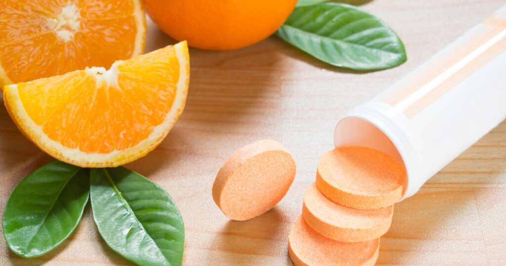 Vitamin c effervescent tablet spilling out of white plastic bottle with fresh juicy orange fruits and green leaf on wood table. Vitamins from foods or supplements choices. Health and medical concept.