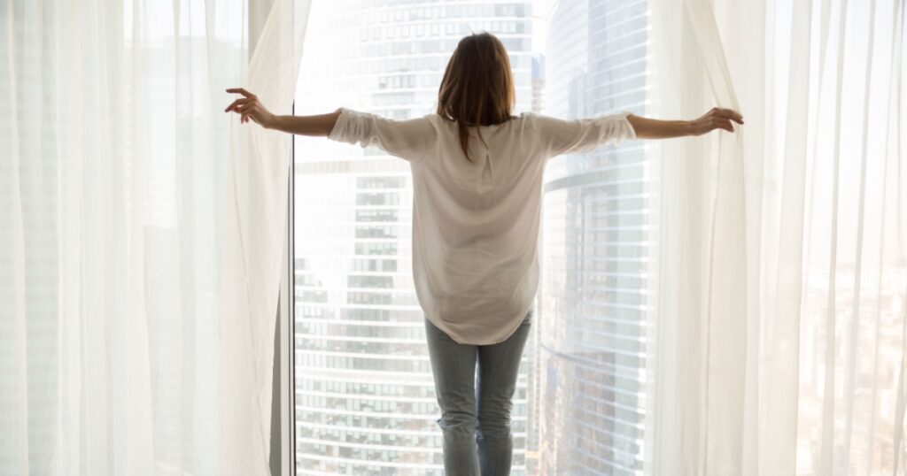 Rear view at rich woman standing looking out of full-length window of luxury modern apartment or hotel room opening curtains in the morning enjoying sunlight and city skyscrapers view feeling happy