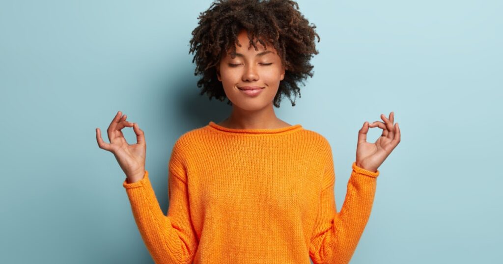 Mindful peaceful Afro American woman meditates indoor, keeps hands in mudra gesture, has eyes closed, tries to relax after long hours of working, holds fingers in yoga sign, isolated on blue wall