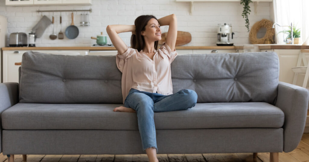 In cozy living room happy woman put hands behind head sitting leaned on couch 30s european female enjoy lazy weekend or vacation, housewife relaxing feels satisfied accomplish chores housework concept