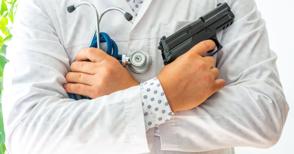 Doctor stands in full height with his arms folded on his breasts and holds gun in one palm and phonendoscope in other. Concept photo of a doctor killer or a bad specialist in medical practice