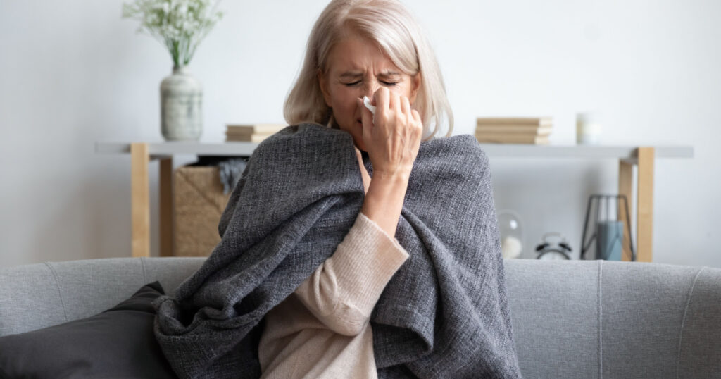 Middle-aged 50s sick frozen woman seated on sofa in living room covered with warm plaid sneezing holding paper napkin blow out runny nose feels unhealthy, seasonal cold, weakened immune system concept