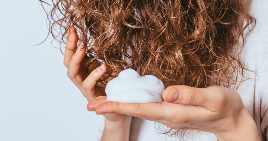 Woman's hands apply styling mousse to her curly hair on white studio background, close-up.