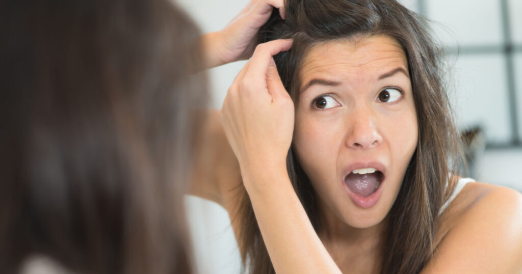 Horrified young woman looking in the bathroom mirror staring open mouthed at the first grey hair on her scalp, a first sign of ageing, or noticing that she is suffering from dandruff