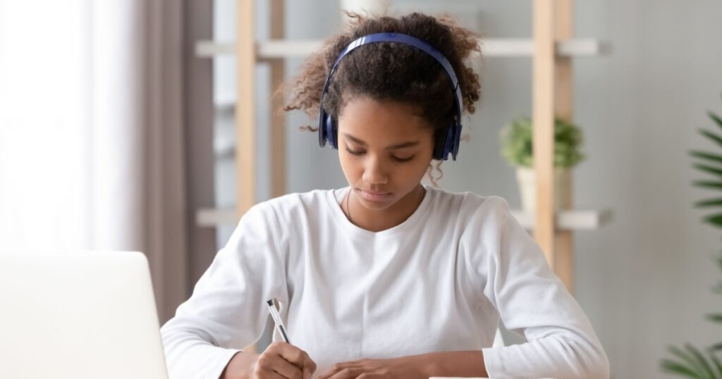 Focused african american teenage girl wearing headphones writing notes study with laptop and books, serious black female high school teen student listening audio course or music while doing homework