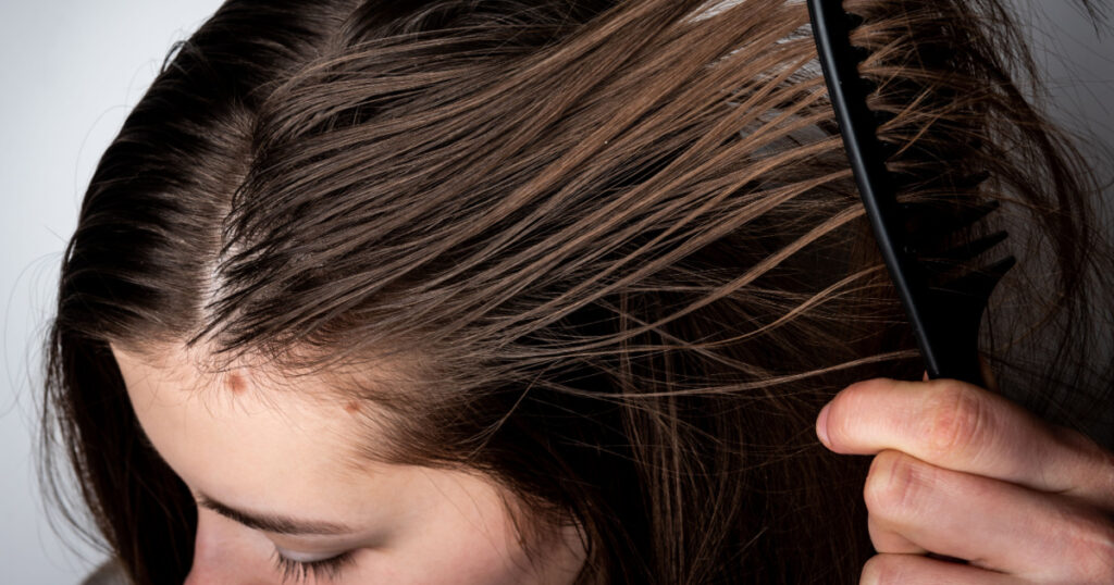 Young woman combing dirty greasy hair on gray background.