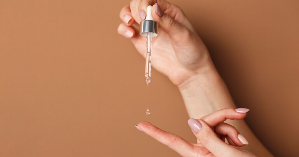 Hands of cropped white woman holding cosmetic serum pipette on the orange background.