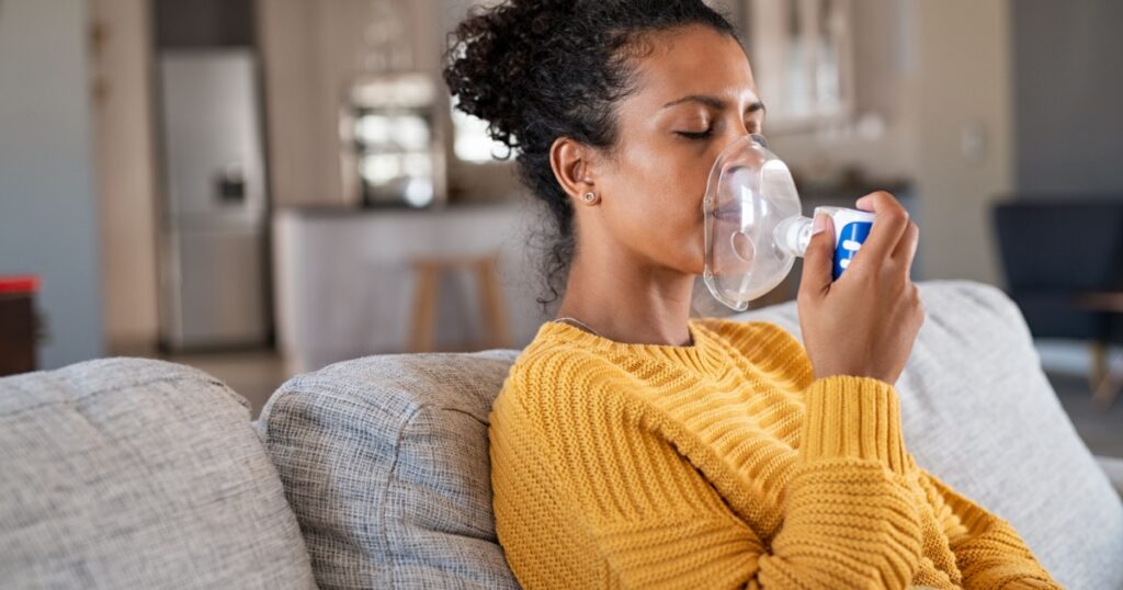 Black woman holding a mask nebulizer inhaling fumes medication into lungs. African sick lady inhaling through inhaler mask at home. Self treatment of the respiratory tract using inhalation nebulizer.