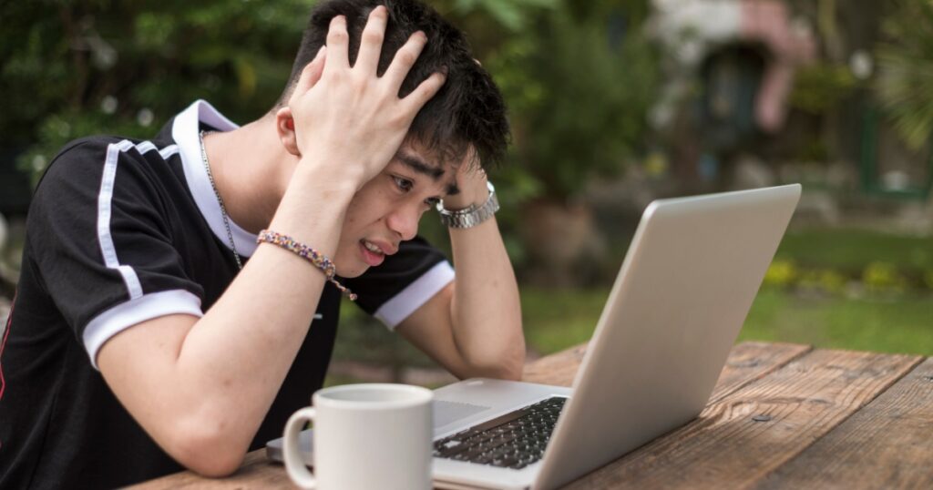 A young asian man is extremely frustrated at his workload while sitting at a table at a garden area outside the house or a park. Concept of stress during studying or working.