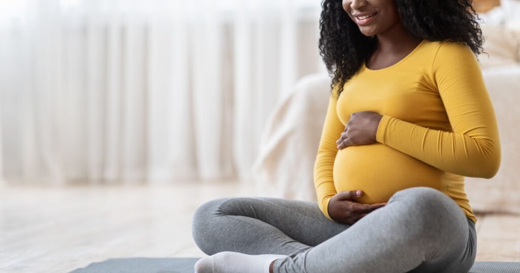 Pregnant african american woman sitting on yoga mat in living room, hugging her belly, copy space. Unrecognizable black expecting lady doing sport at home. Healthy lifestyle during pregnancy