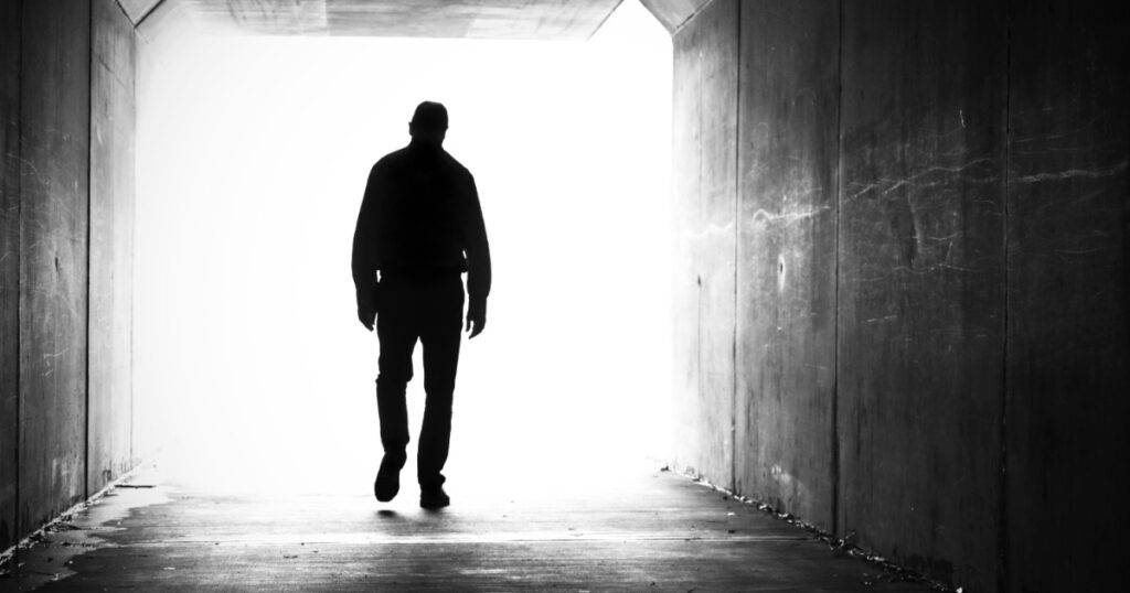 silhouette of man, shoulders slumped walking out of a dark tunnel towards a bright light.