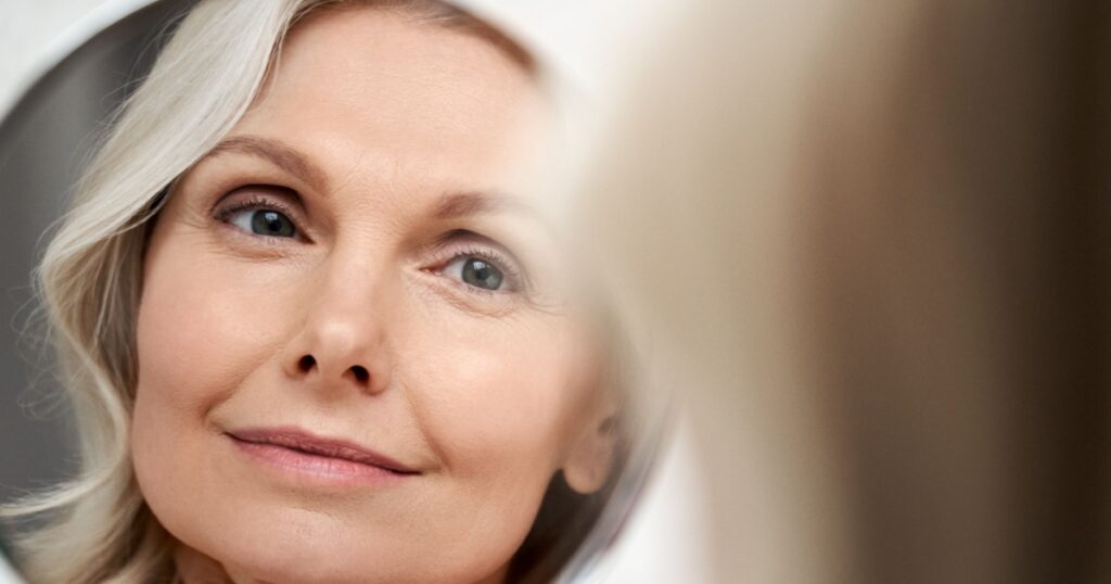 Happy 50s middle aged woman model touching face skin looking in mirror reflection. Smiling mature old lady pampering, healthy moisturized skin care, aging beauty, skincare treatment
