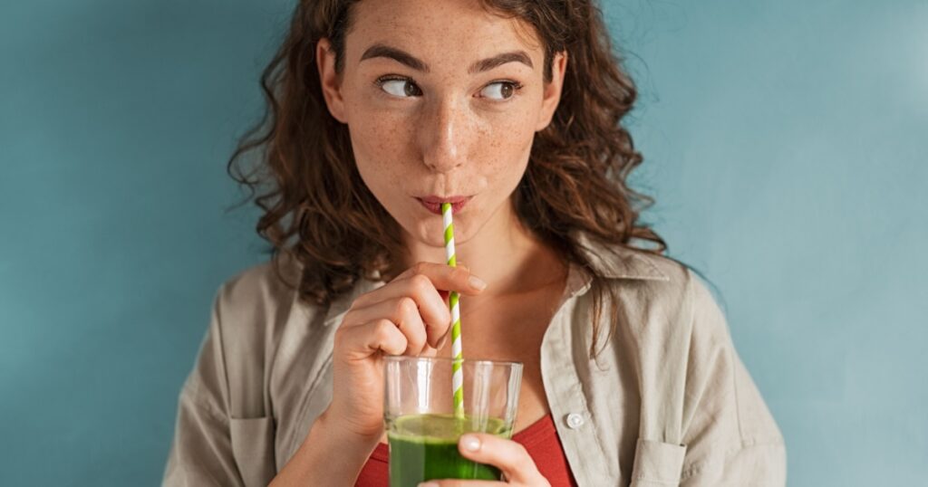 Beautiful woman drinking an organic green smoothie. Fit young woman drinking detox juice using paper straw isolated against blue background. Healthy girl enjoy detox drink and looking away.
