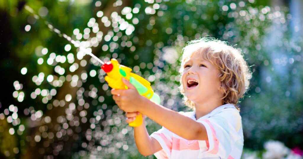 Kids play with water gun toy in garden. Outdoor summer fun. Little boy playing with water hose in sunny backyard. Party game for children. Healthy activity for hot sunny day.