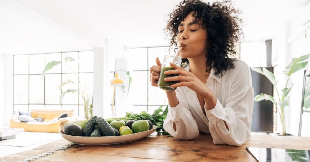 Young african american woman drinking green juice with reusable bamboo straw in loft apartment. Home concept. Healthy lifestyle concept. Copy space