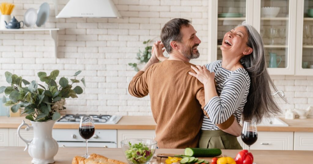 Happy cheerful middle-aged mature couple family parents dancing together in the kitchen, preparing cooking food meal for romantic dinner, spending time together. Active seniors
