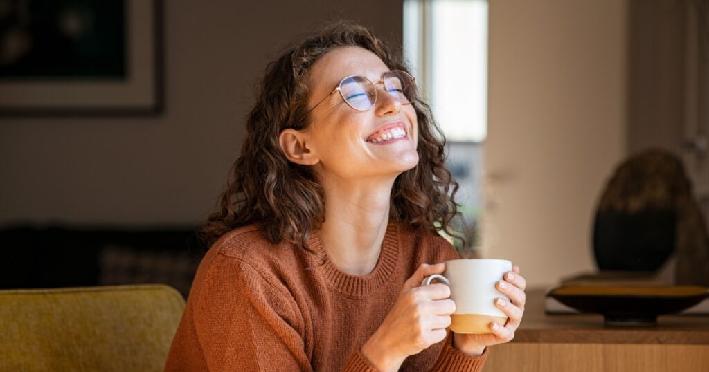Portrait of joyful young woman enjoying a cup of coffee at home. Smiling pretty girl drinking hot tea in winter. Excited woman wearing spectacles and sweater and laughing in an autumn day.
