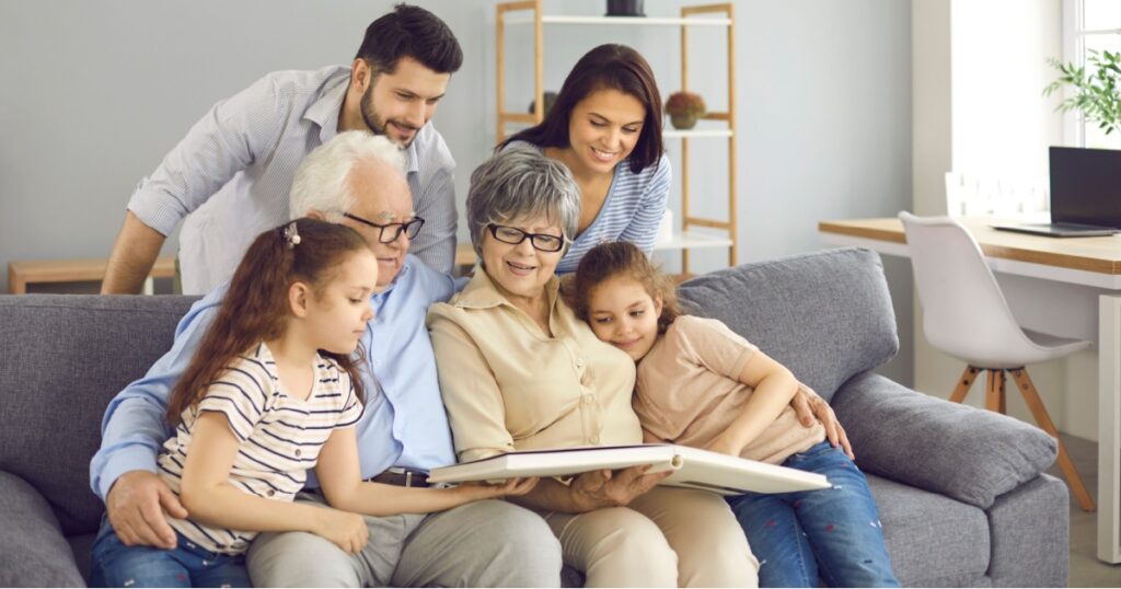 Happy extended family reading together. Young parents, little kids and mature grandparents sitting on sofa at home, enjoying book of good kind educational stories or looking through family photo album