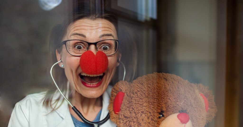 Portrait of a smiling female doctor with red clown nose holding teddy bear, looking at camera.