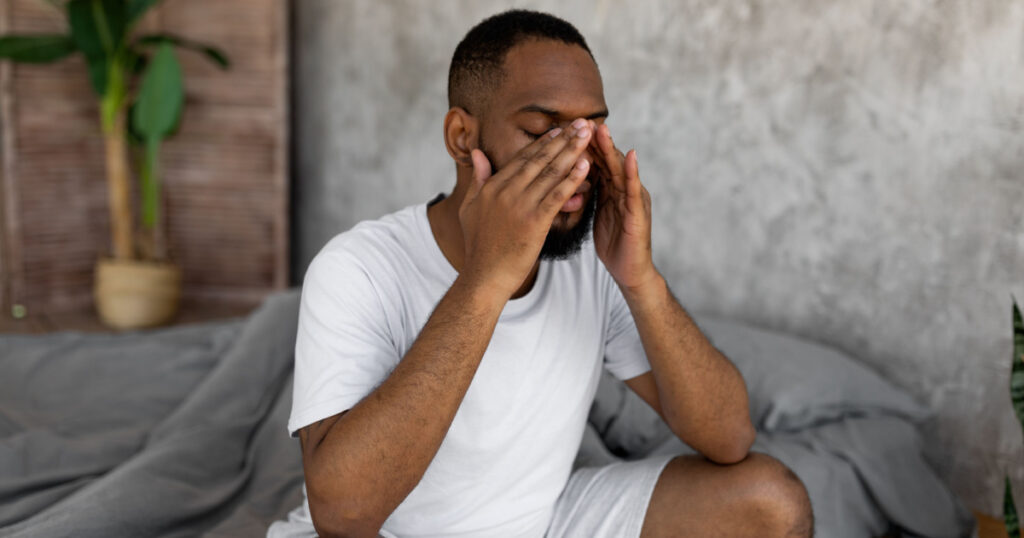 Young black man suffering from headache or migraine, rubbing eyes after waking up. Stressed guy sitting on bed with painful face expression feeling terrible hangover, unpleasant weakness or depression