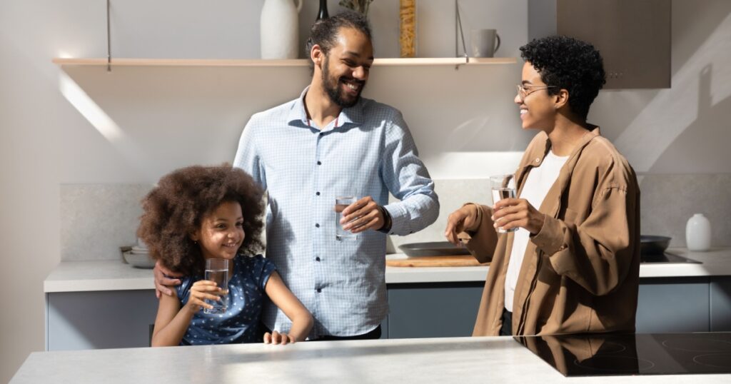 Happy laughing African American family couple and healthy daughter kid drinking clean still pure water in home kitchen, holding glasses, talking, laughing, keeping healthy hydration