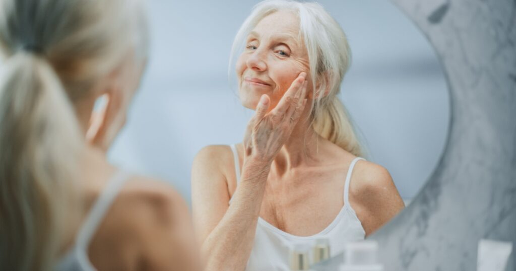 Beautiful Senior Woman Morning Bathroom Routine, Looks into Mirror Touches Her Face with Sensual Movement of Hands. Elderly Female Natural Beauty. Dignity and Grace in Old Age. Over the Shoulder Shot.