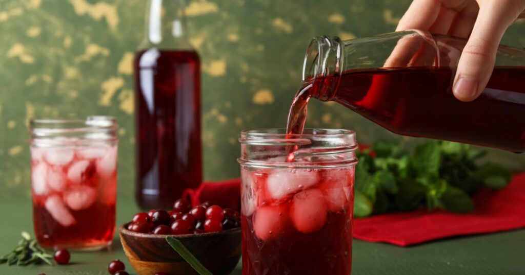 Woman pouring healthy cranberry juice from bottle into glass on color wooden background