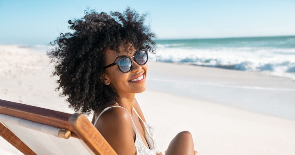 Portrait of happy young black woman relaxing on wooden deck chair at tropical beach while looking at camera wearing spectacles. Smiling african american girl with fashion sunglasses enjoying