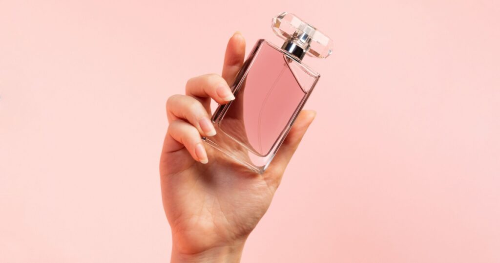 A template for perfume and toilet water. A woman's hand elegantly holds a glass perfume bottle on a pink background, close-up. Copy space. The concept of perfumery and beauty.