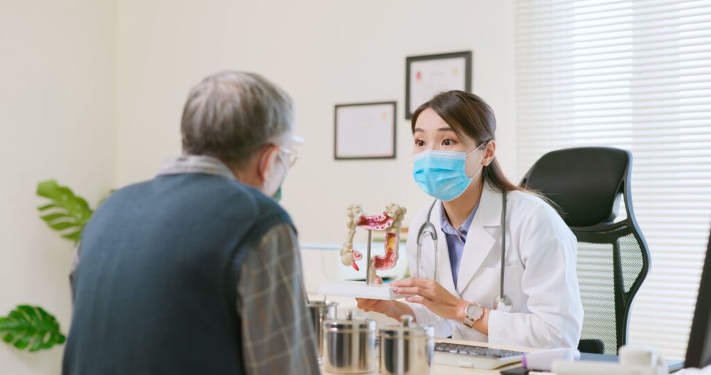 asian female doctor wearing face mask is showing a model of the large intestine and explaining to elder senior man patient in hospital - ok gesture