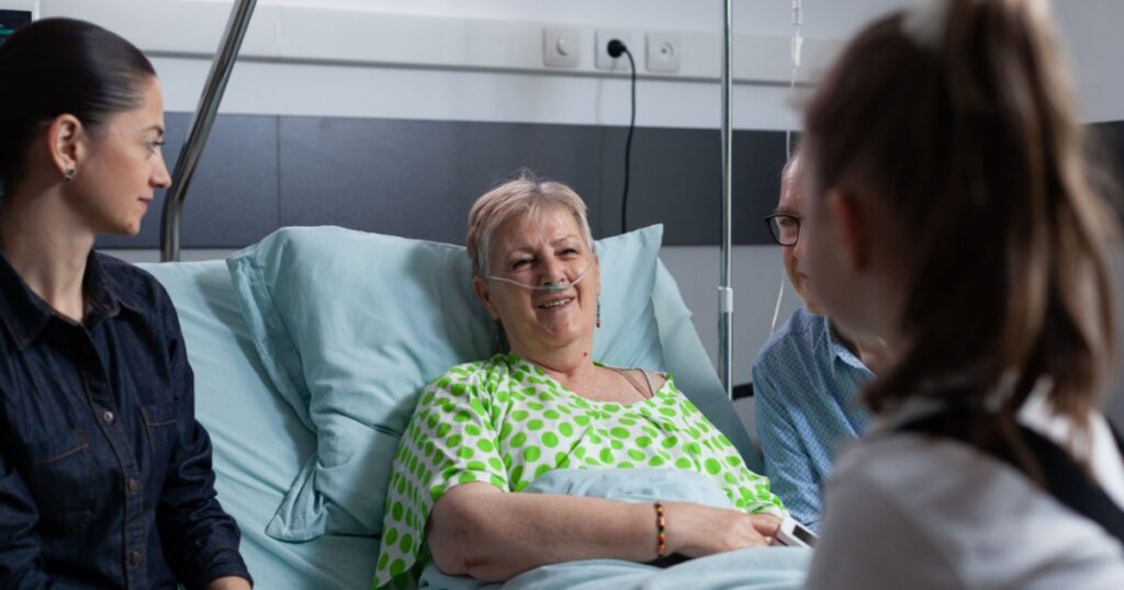 Smiling elderly woman under medical observation in hospital, chatting happily with girl. Relatives visiting senior lady during hospital stay. Happy older female patient with several people at