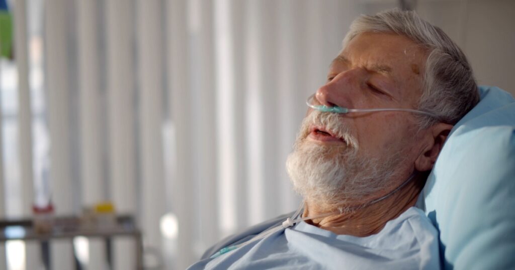 Close up portrait of senior man with nasal breathing tube lying in hospital bed. Ill aged male patient with oxygen cannula breathing heavily having respiratory disease