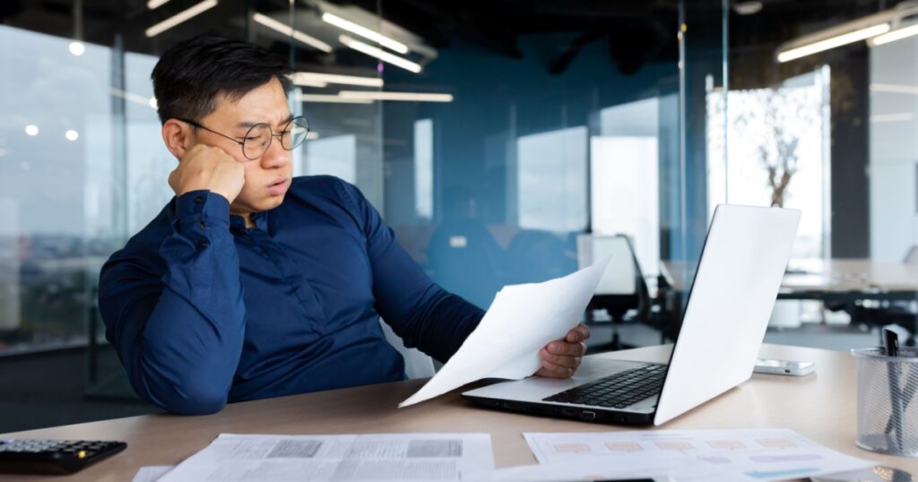 Tired young Asian man, accountant, businessman, freelancer. He sits in the office at a table with a laptop, looks through documents, holds his head in his hands, sighs a hard and boring day.