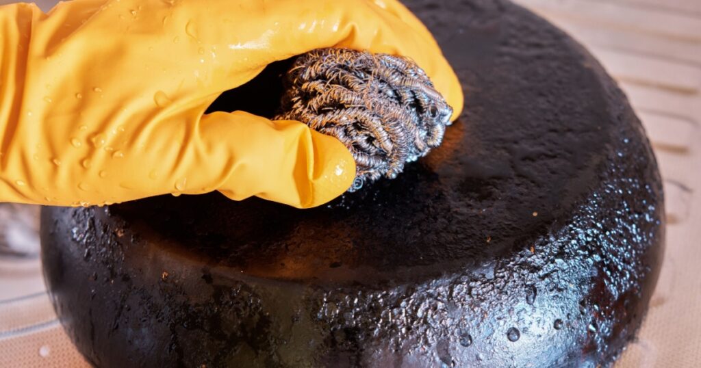 A man's hand in a protective glove cleans a pan with a burnt bottom using a metal kitchen sponge.
