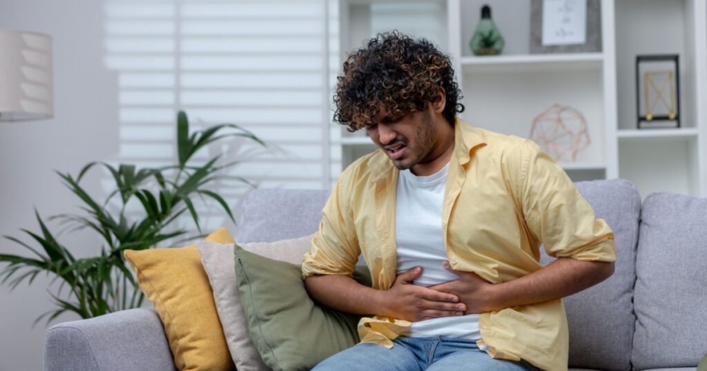 Young man sick at home sitting on sofa, Latin American has severe stomach pain and nausea, poisoned by poor quality food.