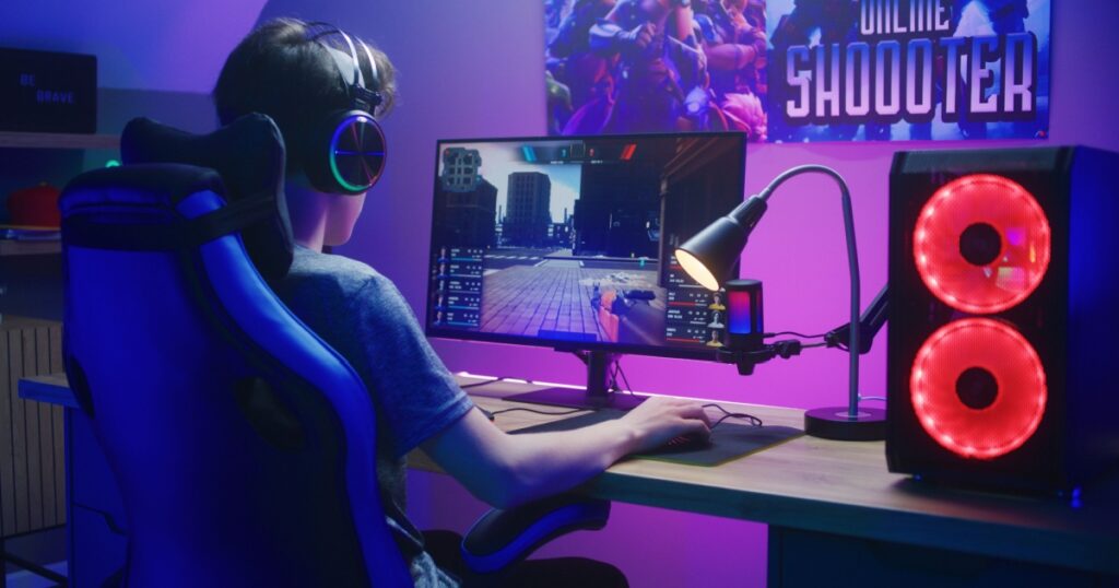 Young gamer in headphones plays in first person shooter on computer. Online video game live streaming or esports tournament. Desk illuminated by RGB LED strip light. Gaming at home concept.