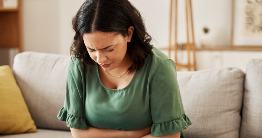 Stress, stomach pain and woman on a sofa with menstruation, gas or constipation, pms or nausea at home. Gut health, anxiety and lady with tummy ache in living room from ibs, bloated or endometriosis