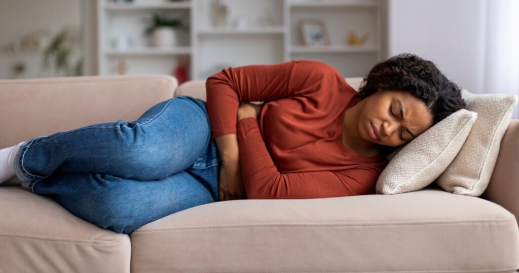 Young black woman clutching her abdomen in discomfort at home, african american female suffering from menstrual pain or stomach issues, lying on sofa in her living room, free space
