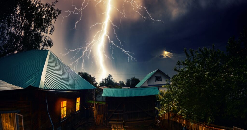 Thunderbolt over the house with dark stormy sky on the background and moon shining through the cloud