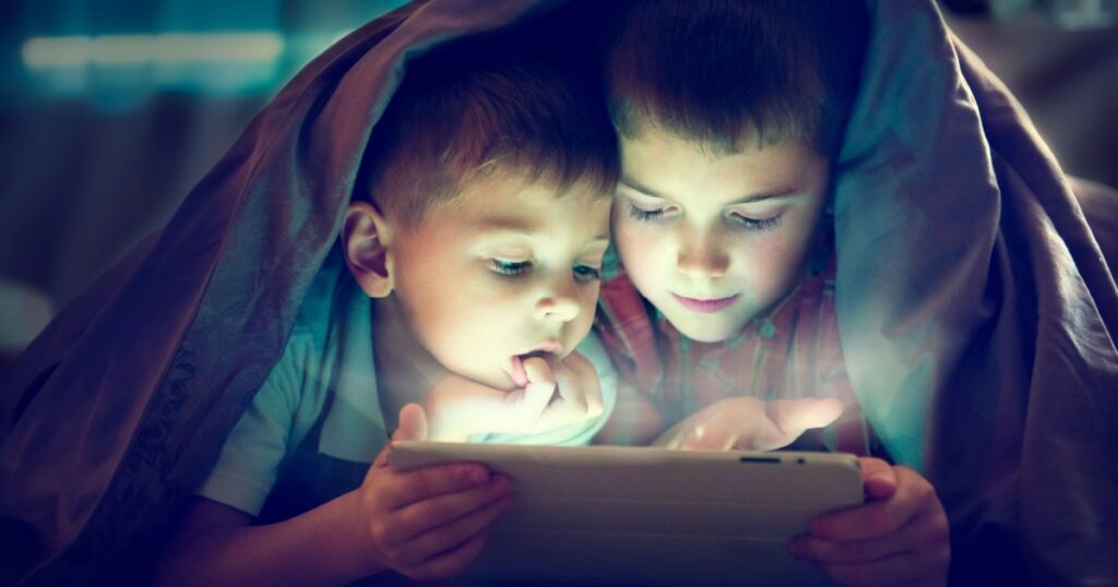 Two kids using tablet pc under blanket at night. Brothers with tablet computer in a dark room