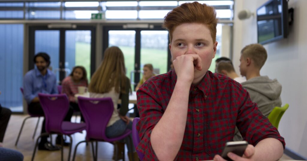 Stressed schoolboy sitting away from people at school. He has a smartphone in his hand and is looking away with his hand to his face.