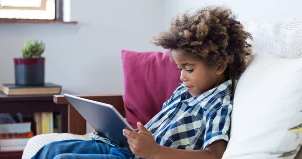 Little african boy sitting on sofa and playing game on digital tablet. Portrait of a young black child at home watching cartoon on the laptop. Modern kid and education technology.