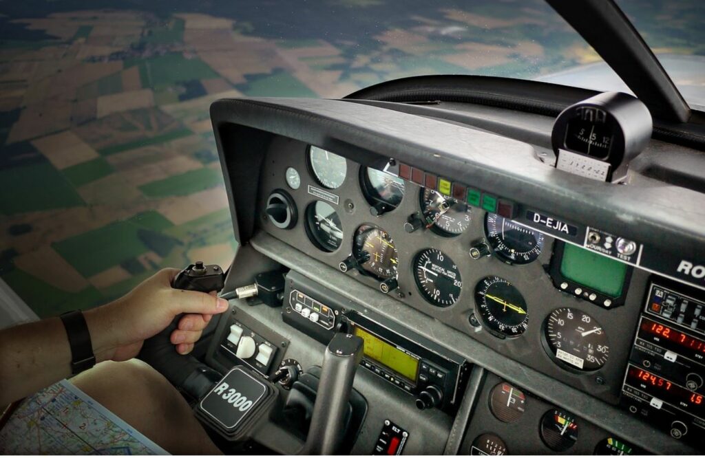 Pilot hand and airplane cockpit, green and tan land in the background.