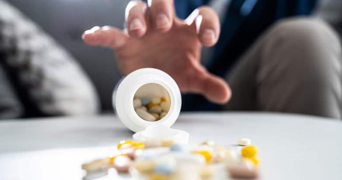 bottle of pills spilling over with contents pouring onto table. A hand is reaching for the bottle.