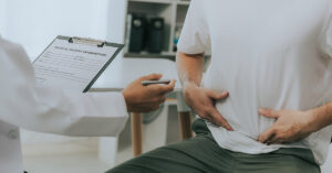 7 Signs Your Stomach Trouble Might Be Crohn’s Disease person holding their abdomen while seeing doctor. Crohn's Disease symptoms concept